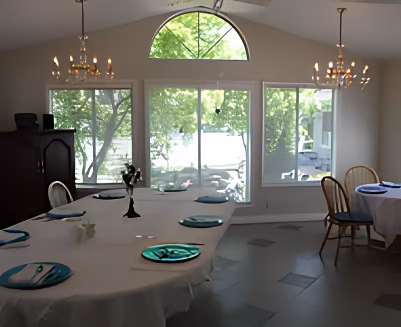 Dining room with a large oval table covered with a white tablecloth, set with blue plates and napkins. There are wooden chairs around the table, two chandeliers hanging from the ceiling, and large windows showing green trees outside.