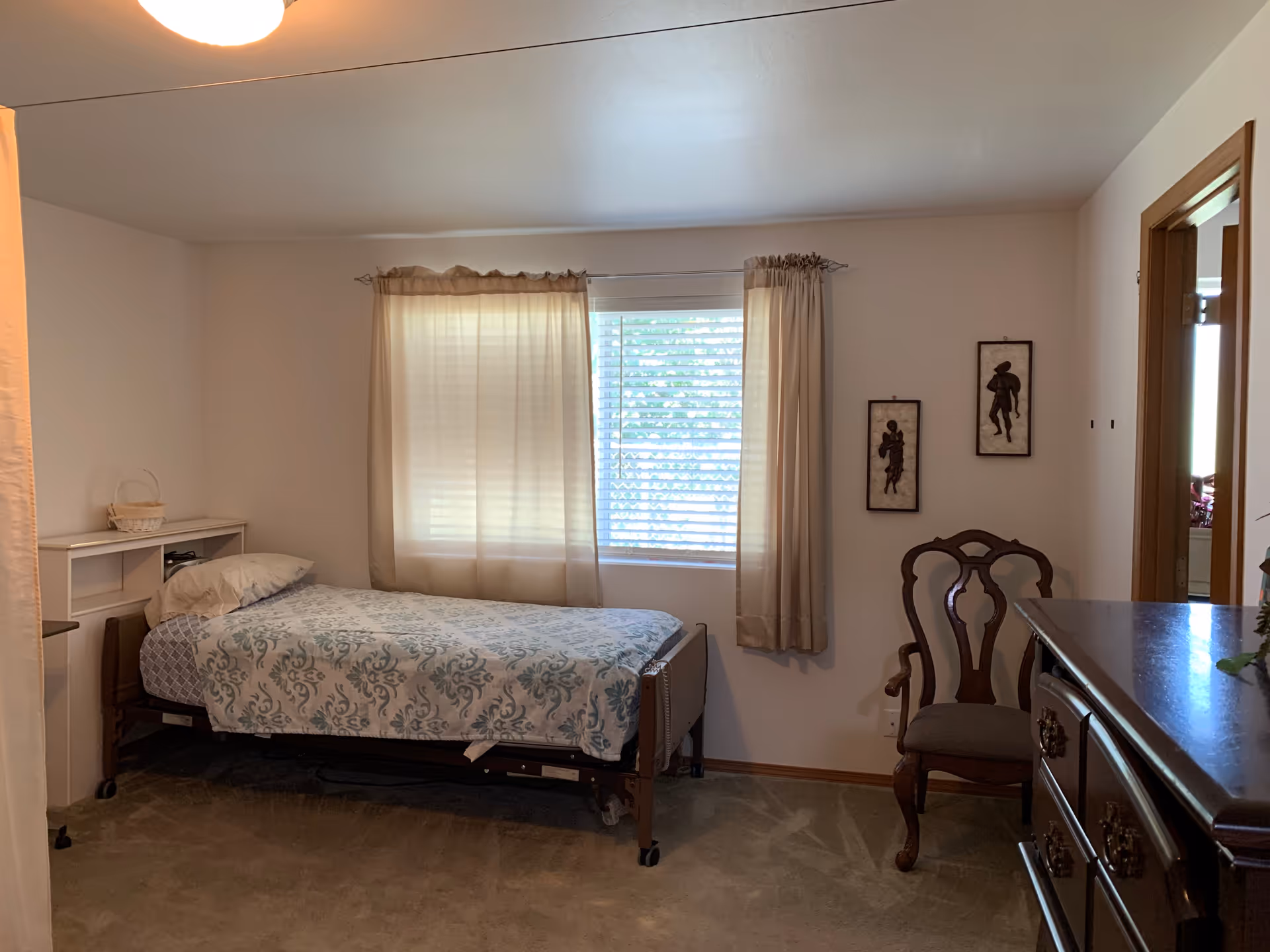 Furnished bedroom with a single bed on a wheeled frame beneath a curtained window, a wooden chair and a dresser.