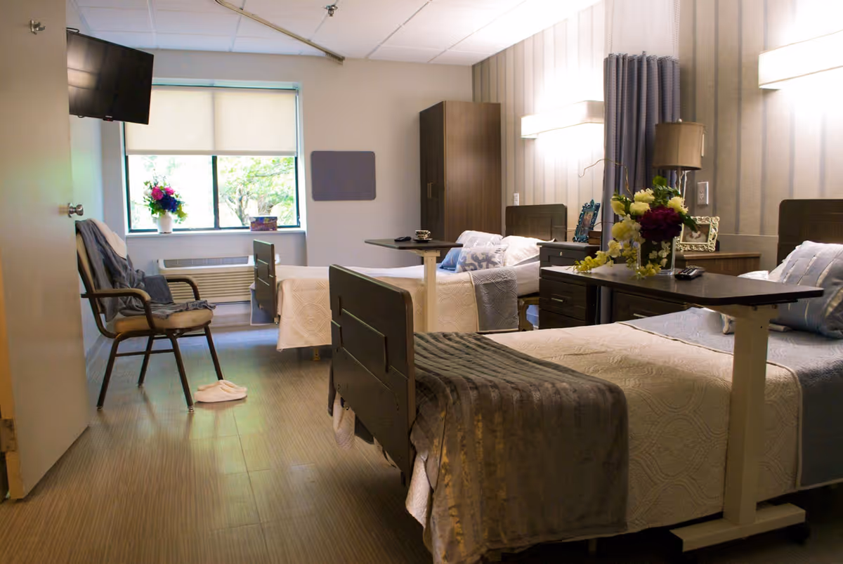 A well-lit nursing center room with two hospital-style beds, each with white bedding and gray blankets. There are overbed tables with flower arrangements on them, a chair with a blanket draped over it, a wall-mounted TV, and a window with a view of greenery outside. The room has a calm and clean atmosphere with soft lighting and neutral-colored walls.