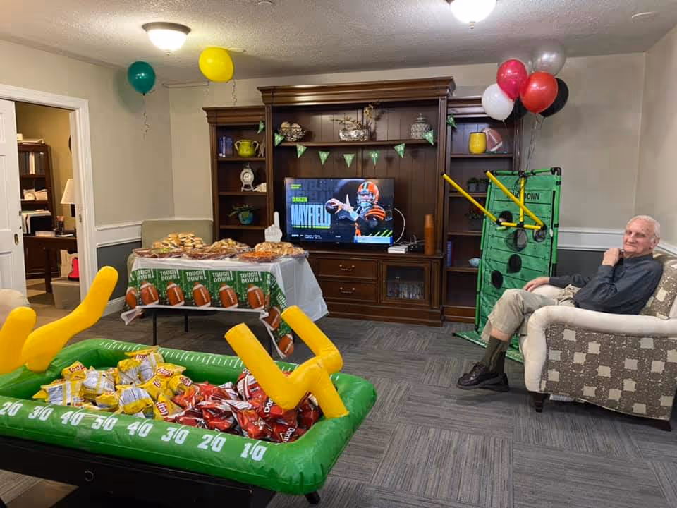 A senior man sitting on a patterned armchair in a decorated living room with a football theme. There is a wooden entertainment center with a TV displaying a football player, a table with snacks and sandwiches covered with a football-themed tablecloth, balloons in green, yellow, red, white, and black colors, and an inflatable football goalpost filled with bags of chips.