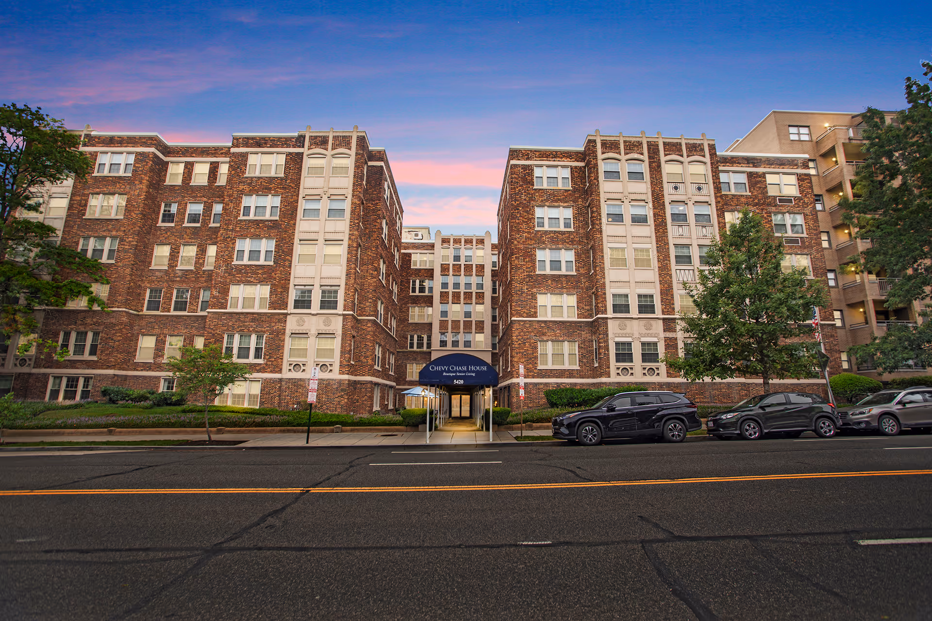 Front exterior view of a multi-story brick building named Chevy Chase House with a blue awning over the entrance, several parked cars along the street, and trees on either side under a colorful sunset sky.