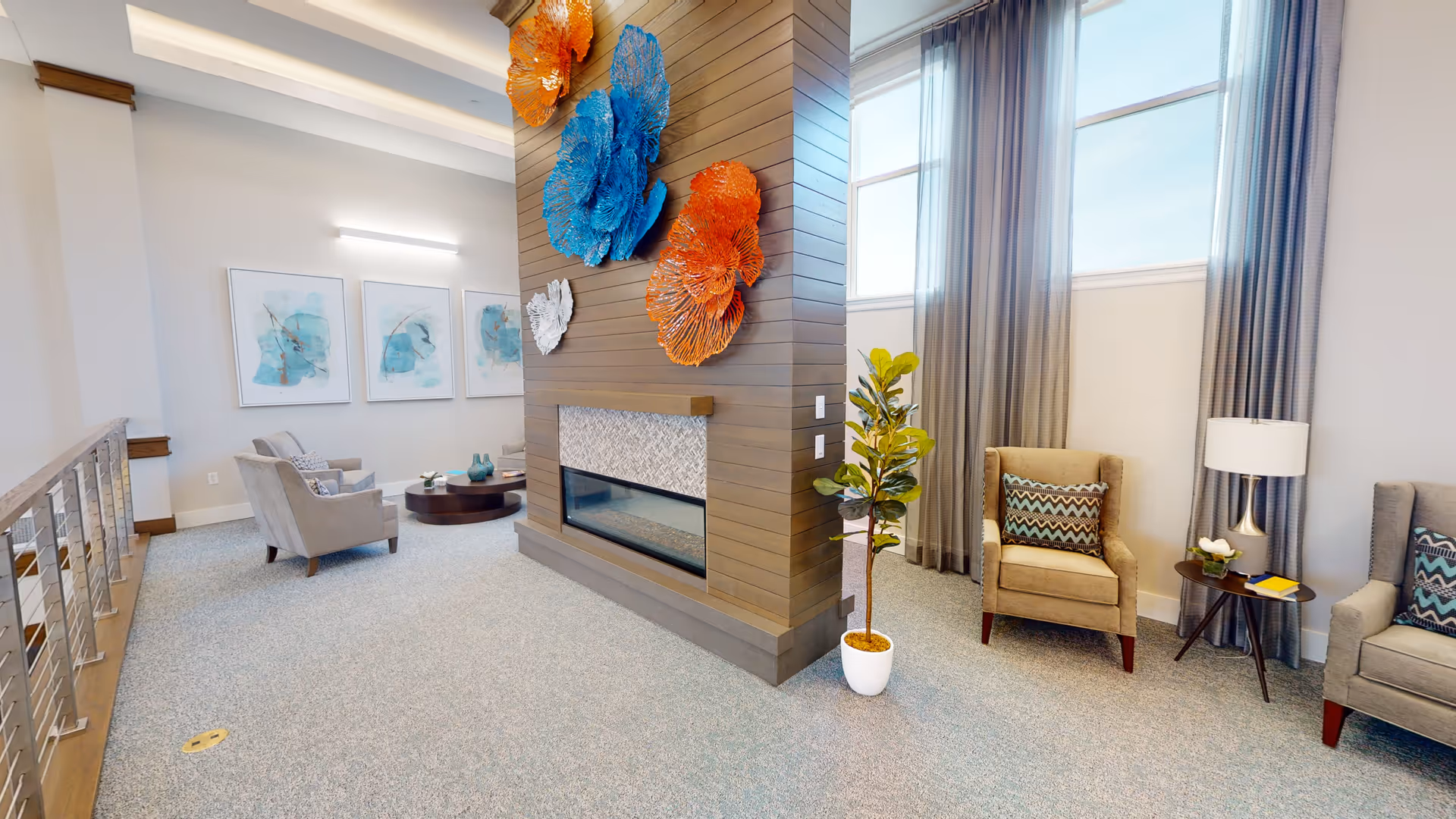 Bright communal lounge with armchairs, a central wood-paneled fireplace wall adorned with blue and orange sculptures, and tall windows with curtains.