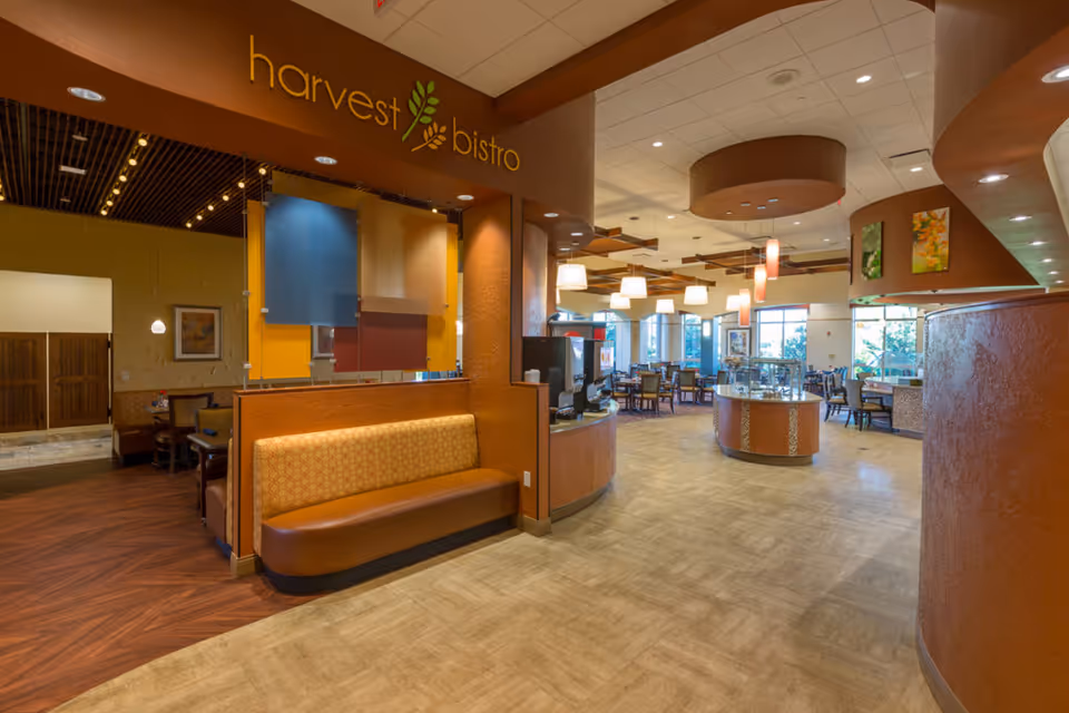 Interior view of a dining area named 'harvest bistro' with warm lighting, wooden and cushioned seating, tables and chairs arranged for dining, and large windows letting in natural light.
