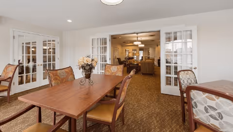 A dining room with wooden tables and cushioned chairs featuring patterned upholstery. A vase with flowers is placed on the central table. The room has beige walls and carpeted floors. French doors open into a living room area with sofas and warm lighting.