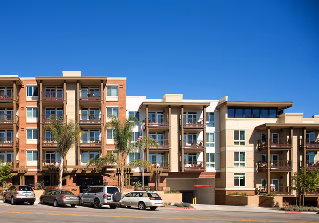 Exterior view of a modern multi-story senior living facility with balconies, palm trees, and parked cars in front under a clear blue sky.