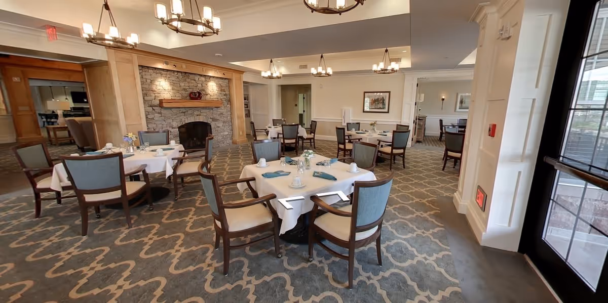 A spacious dining room with several round tables covered with white tablecloths, each set with cups, saucers, and napkins. The room features a patterned carpet, a stone fireplace with a wooden mantle, and multiple chandeliers hanging from the ceiling. There are large windows and a door letting in natural light.