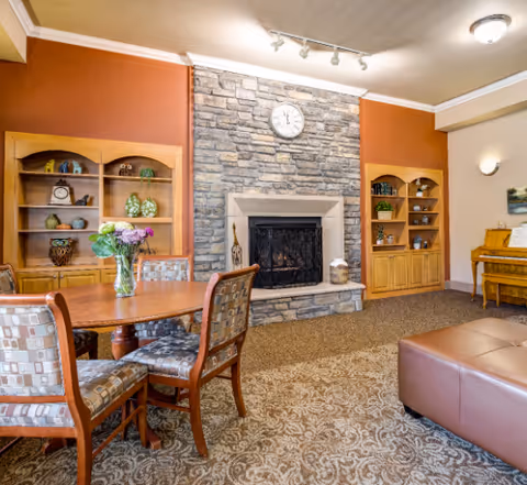 A cozy living room area in an assisted living facility featuring a stone fireplace with a clock above it, two wooden bookshelves with decorative items on either side, a wooden table with four cushioned chairs, a brown leather ottoman, and a piano in the corner. The walls are painted in warm tones with carpeted flooring.