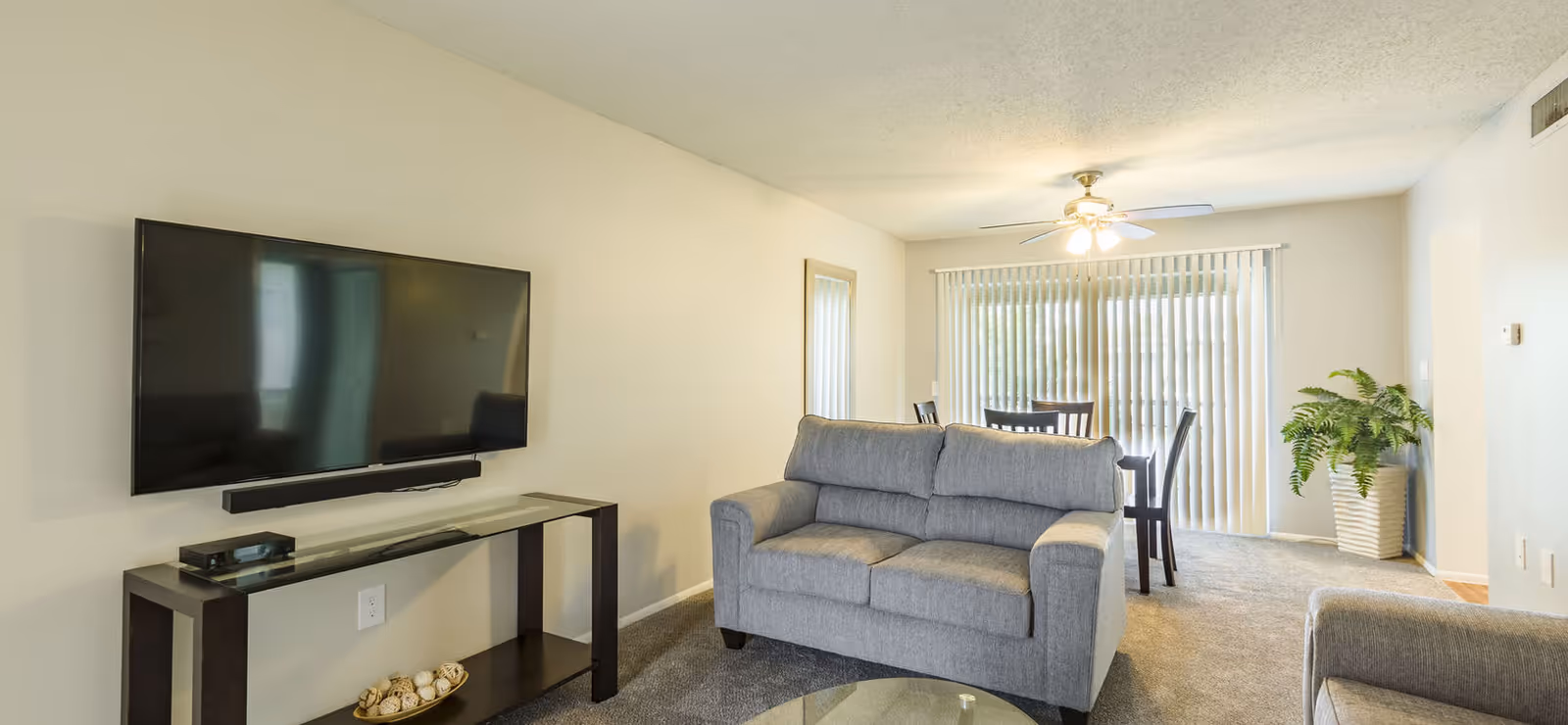 A living room with a gray couch facing a wall-mounted flat-screen TV above a glass and dark wood console table. Behind the couch is a dining area with a wooden table and chairs, a ceiling fan with lights, vertical blinds covering a sliding glass door, and a potted plant in the corner.