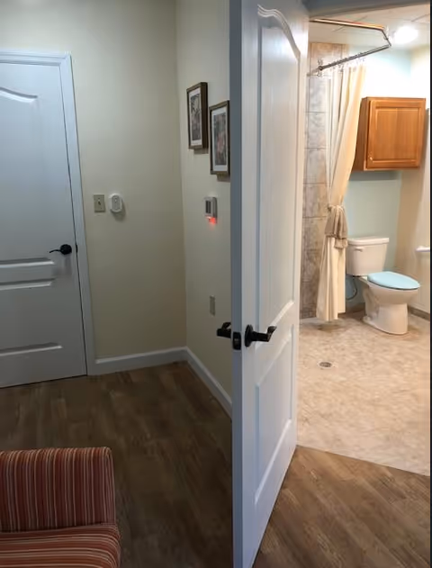 An open door reveals a bathroom with a toilet, shower with curtain and wall cabinet, adjacent to a small living area with a striped chair.