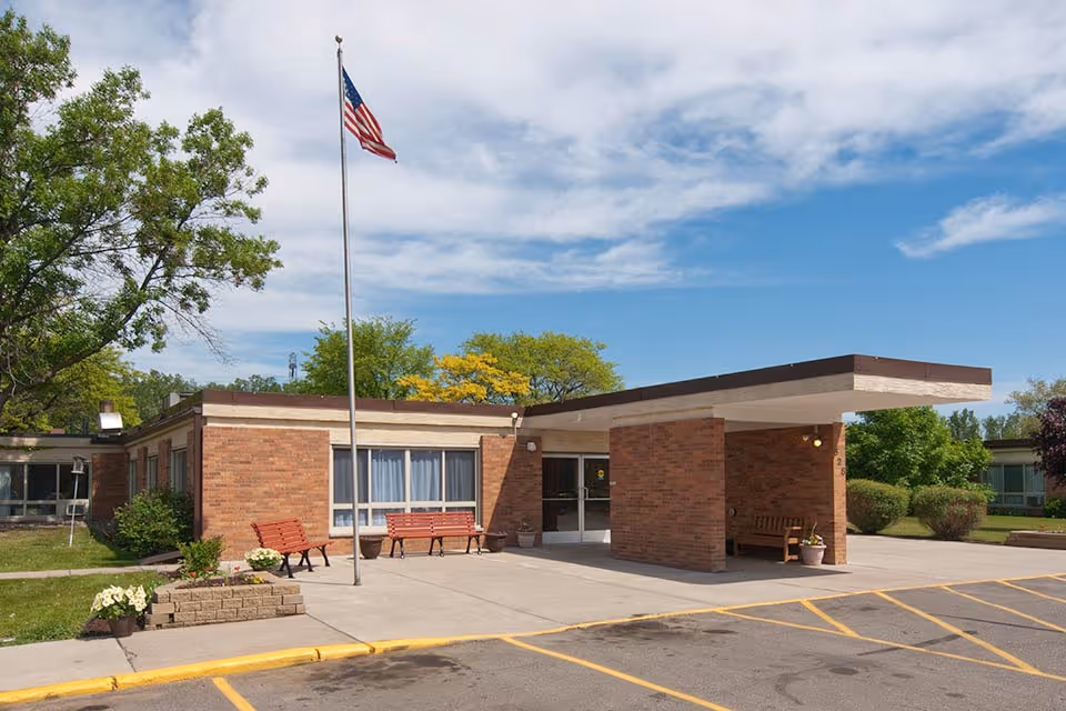 Exterior view of a single-story brick building with a covered entrance, benches outside, an American flag on a flagpole, and surrounding greenery under a partly cloudy blue sky.