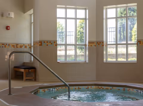 Indoor hot tub or small pool area with tiled walls and floor, two large windows letting in natural light, a metal handrail for entering the water, and a wooden bench with a cushion in the corner.