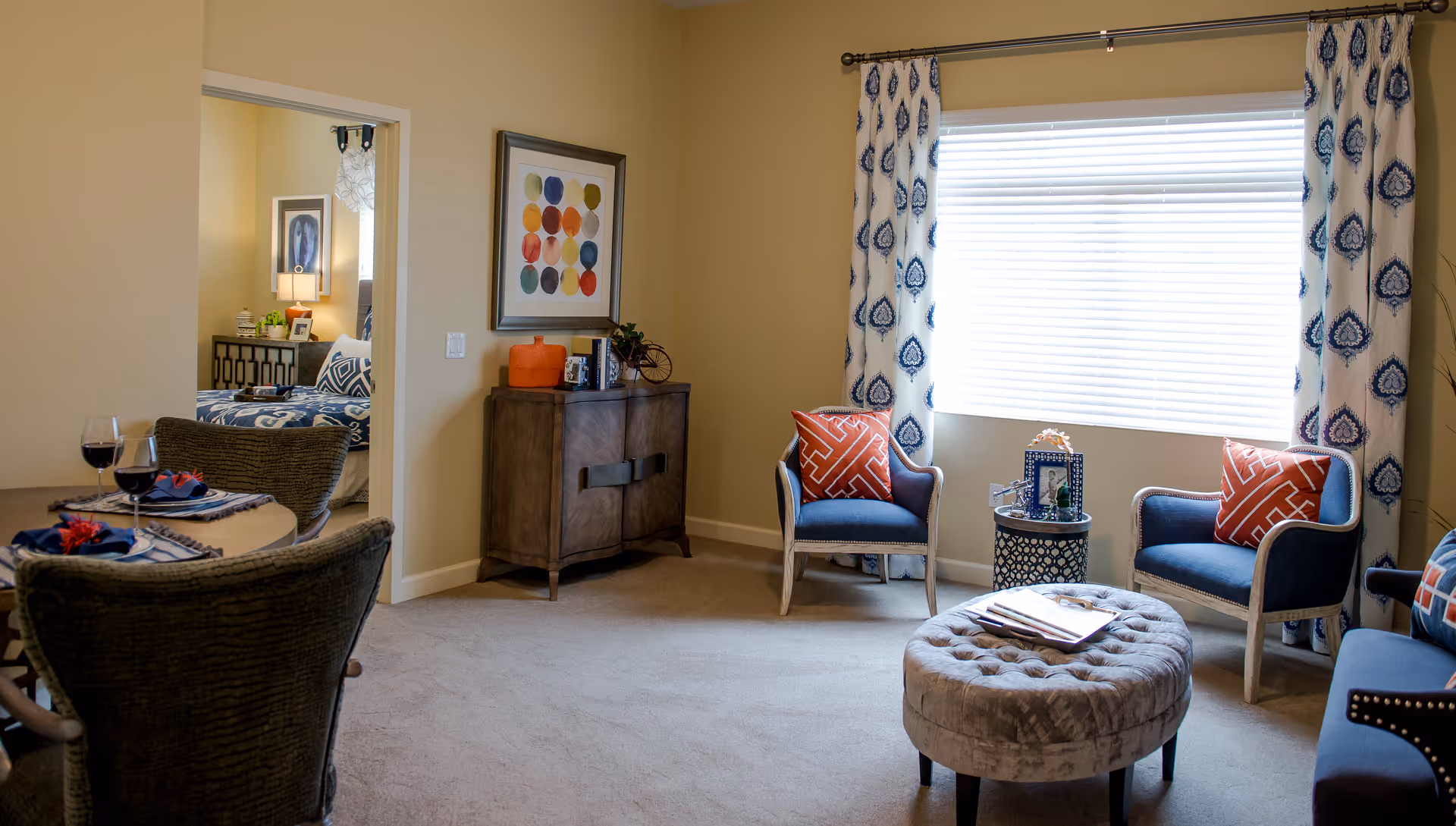 A cozy living room area with two blue armchairs with orange patterned pillows, a round tufted ottoman, a small round side table with a picture frame, and a large window with white blinds and patterned curtains. To the left, there is a small dining table set for two with wine glasses and napkins. A doorway leads to a bedroom with a bed, nightstand, lamp, and framed artwork on the wall.