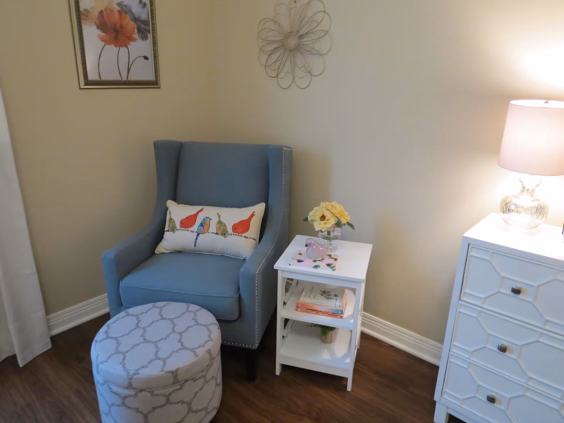 A cozy corner of a room featuring a blue upholstered armchair with a decorative pillow showing birds, a round patterned ottoman, a white side table with books and a vase of yellow flowers, a white chest of drawers with a glass lamp on top, and wall decorations including a framed floral picture and a metal wall art piece.