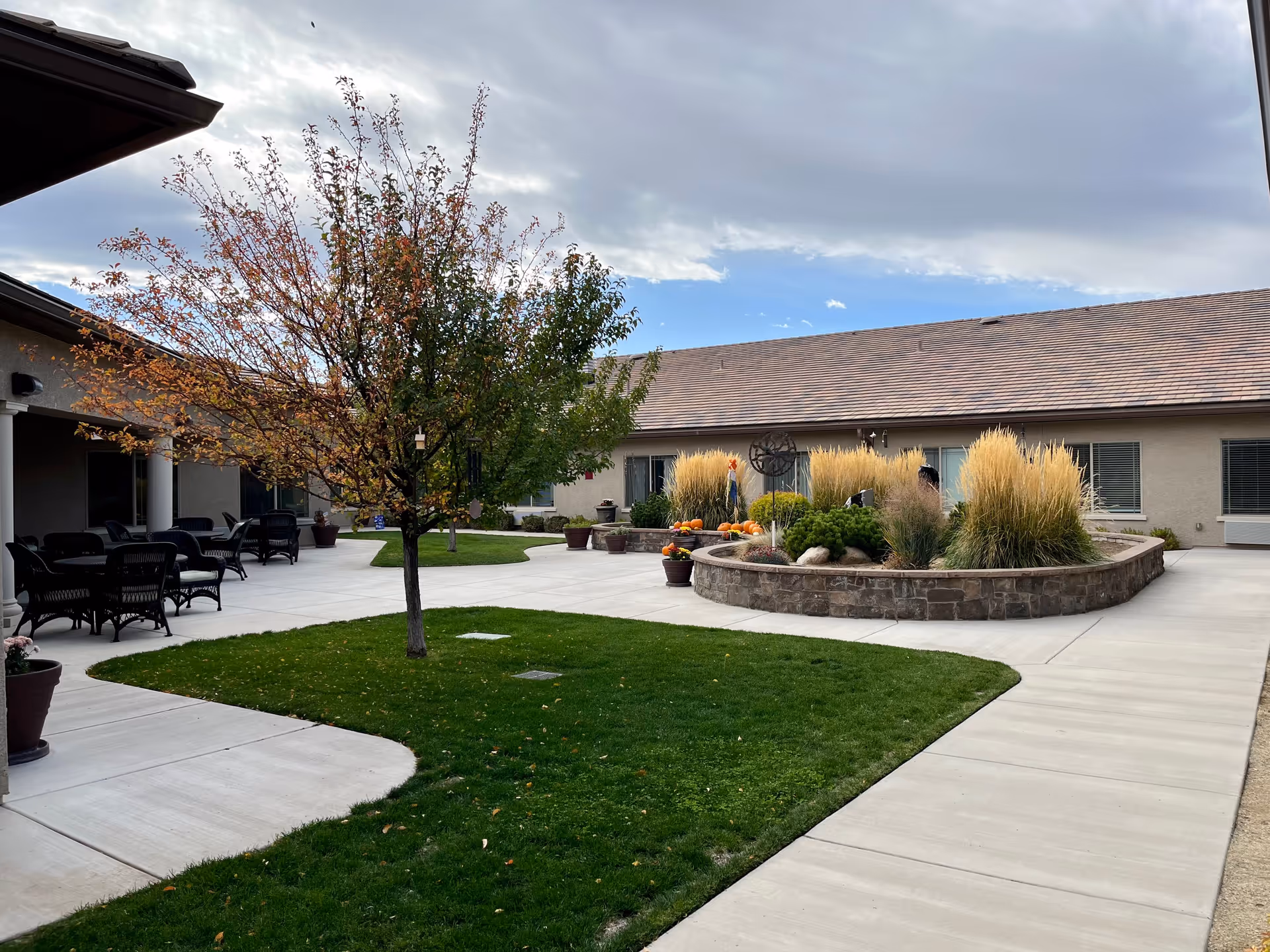 Outdoor courtyard area of Stone Valley Assisted Living & Memory Care featuring a tree with autumn leaves, green grass, paved walkways, a raised stone planter with ornamental grasses and pumpkins, and outdoor seating with black chairs and tables under a covered patio.