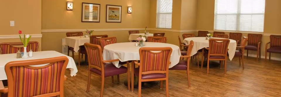 A dining room with several tables covered in white tablecloths, each surrounded by wooden chairs with striped upholstery. The room has wooden flooring, beige walls, framed artwork, and windows with blinds allowing natural light to enter.