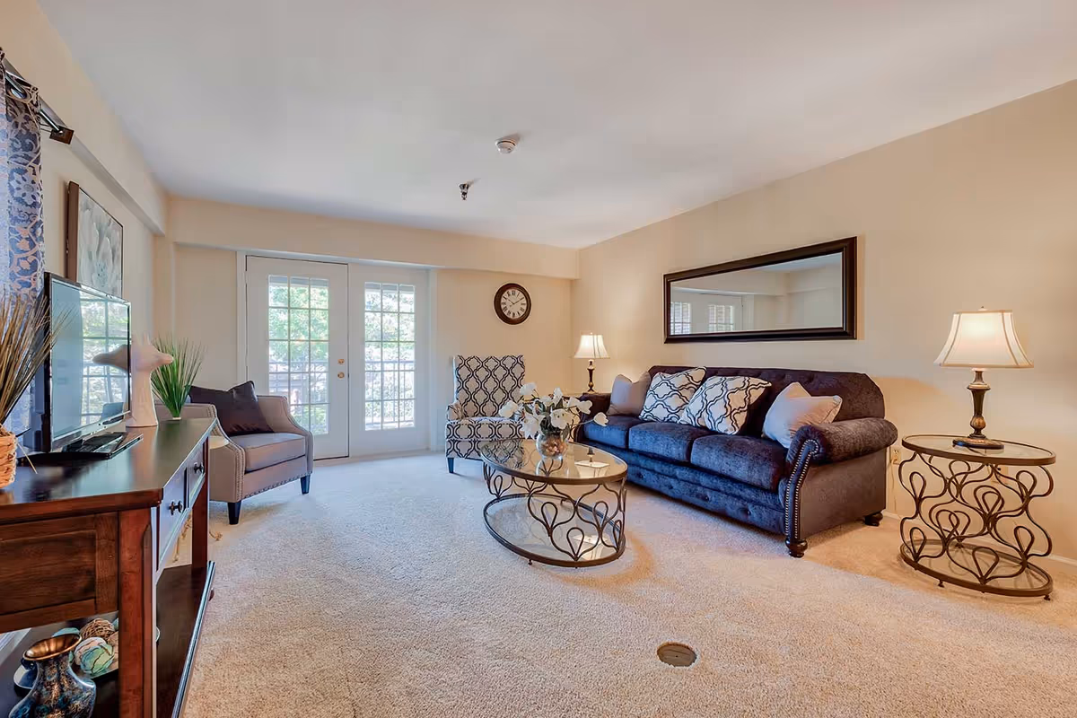 A bright and cozy living room with beige walls and carpet. The room features a dark blue sofa with patterned and solid pillows, a matching patterned armchair, and a gray armchair. There is a glass coffee table with a decorative metal base and a vase with flowers on it. Two side tables with lamps flank the sofa. A large mirror hangs on the wall above the sofa, and a clock is mounted on the wall near French doors that lead outside. A wooden TV stand with a flat-screen TV and decorative items is positioned on the left side of the room.