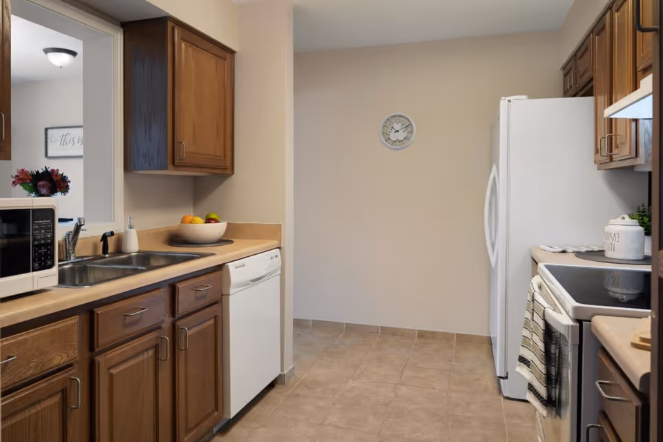 Clean kitchen with wooden cabinets, a sink and dishwasher on the left, refrigerator and stove on the right, and a clock on the back wall.