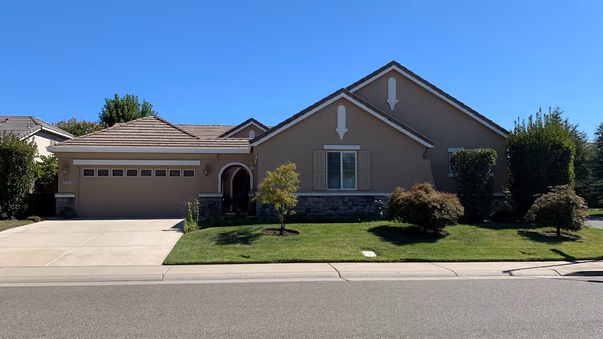 Front exterior view of a single-story residential building with a two-car garage, beige walls, stone accents, and a well-maintained lawn with small trees and shrubs under a clear blue sky.