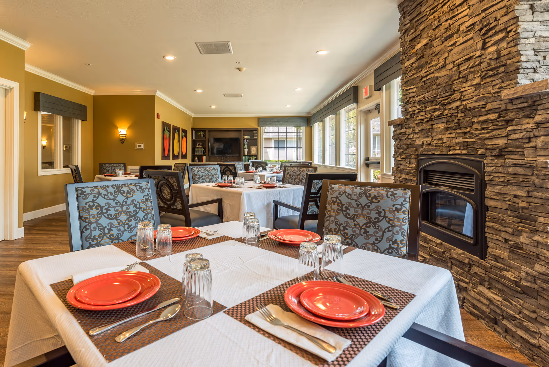 A dining room in a senior living facility with tables set for meals. Each table is covered with a white tablecloth and has red plates, glasses, and silverware. The room features patterned chairs, large windows letting in natural light, a stone fireplace on the right, and colorful artwork on the far wall.