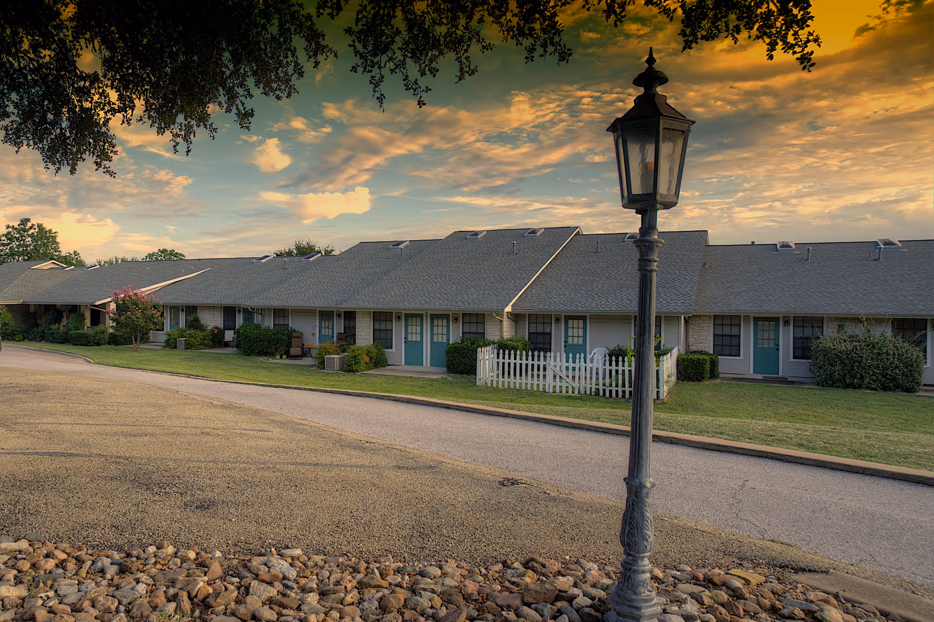 Single-story assisted living building with teal doors, a lamppost and a white picket fence beneath a colorful sunset.