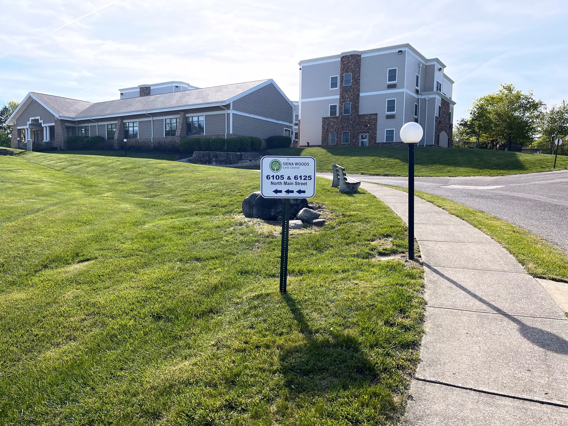 Outdoor view of Siena Woods Care Center showing two buildings with beige and stone exterior walls, a green lawn, a sidewalk, a lamp post, and a sign indicating addresses 6105 & 6125 North Main Street with arrows pointing left.