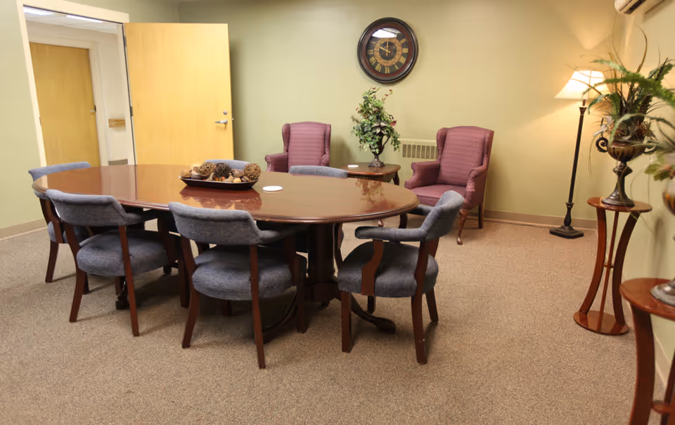 A meeting or dining room with a large oval wooden table surrounded by six blue upholstered chairs. In the background, there are two purple armchairs with a small table between them holding a plant. A round wall clock hangs on the light green wall above a heating vent. A floor lamp and two plant stands with decorative plants are also visible in the room. The door to the room is open, showing a hallway with another closed door.