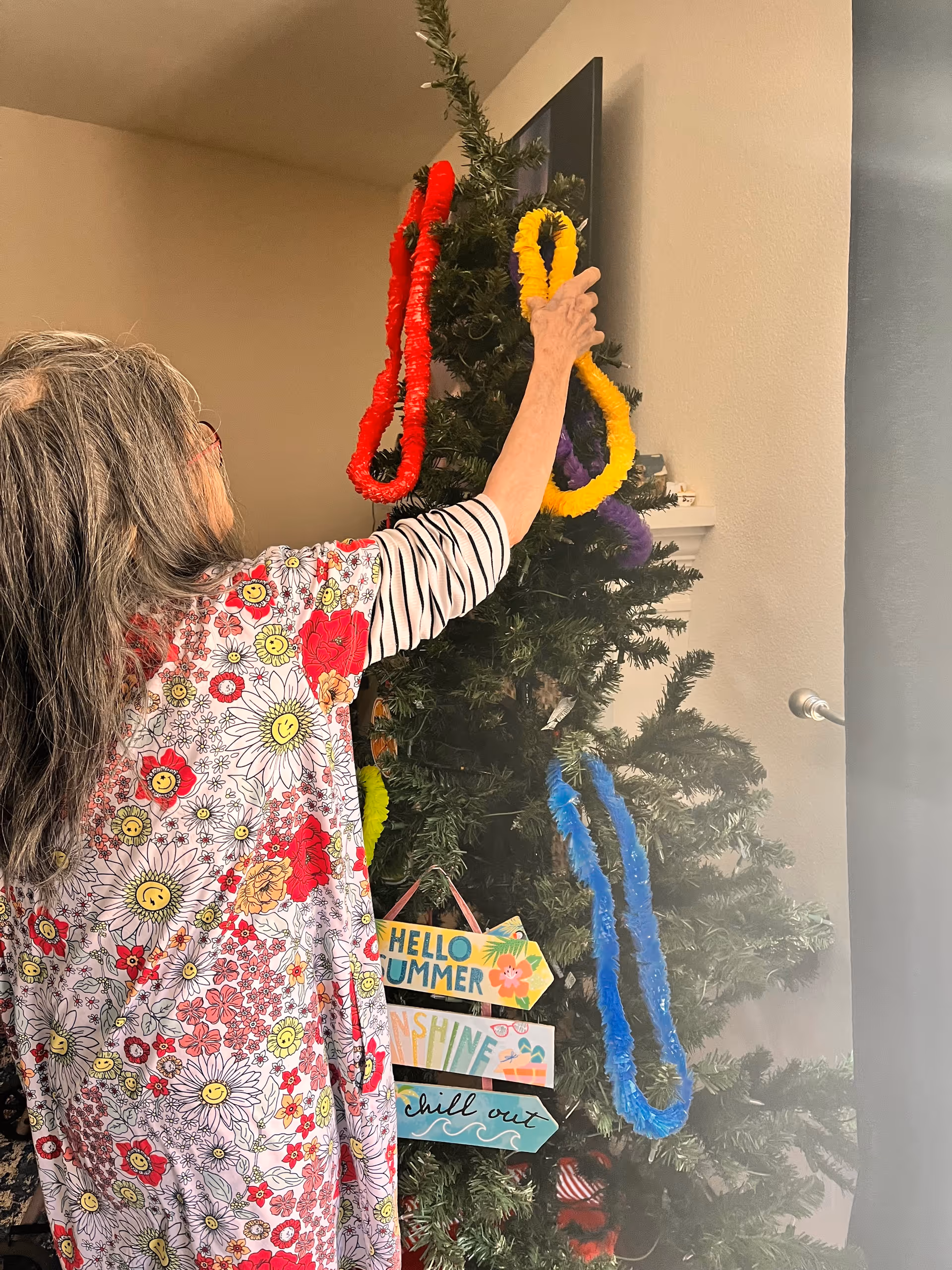 An elderly woman with long gray hair wearing a floral patterned shirt is decorating a Christmas tree with colorful leis in red, yellow, purple, and blue. The tree also has a sign with summer-themed messages such as 'HELLO SUMMER', 'SUNSHINE', and 'chill out'. The setting appears to be indoors with a beige wall and a window with a curtain rod visible.