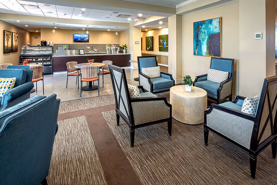 A bright and spacious common area in a senior living facility featuring a seating arrangement with four armchairs around a round table with a small plant. In the background, there are several round tables with chairs, a counter with a display case, and colorful artwork on the walls.