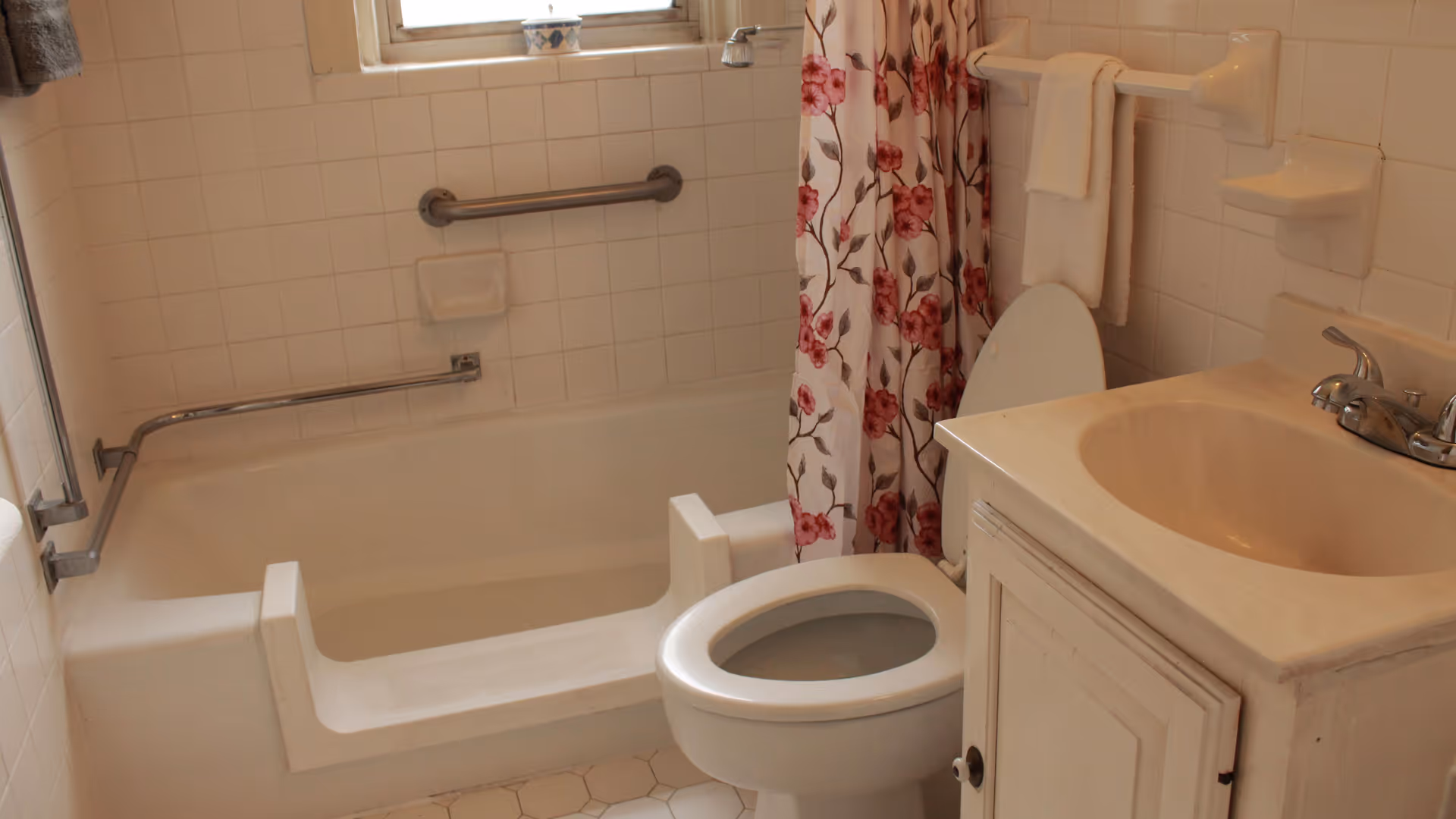 A bathroom with a white bathtub featuring a built-in seat and grab bars, a toilet with the lid open, a sink with a cabinet underneath, and a floral shower curtain. The walls are tiled in white, and there is a window above the bathtub.
