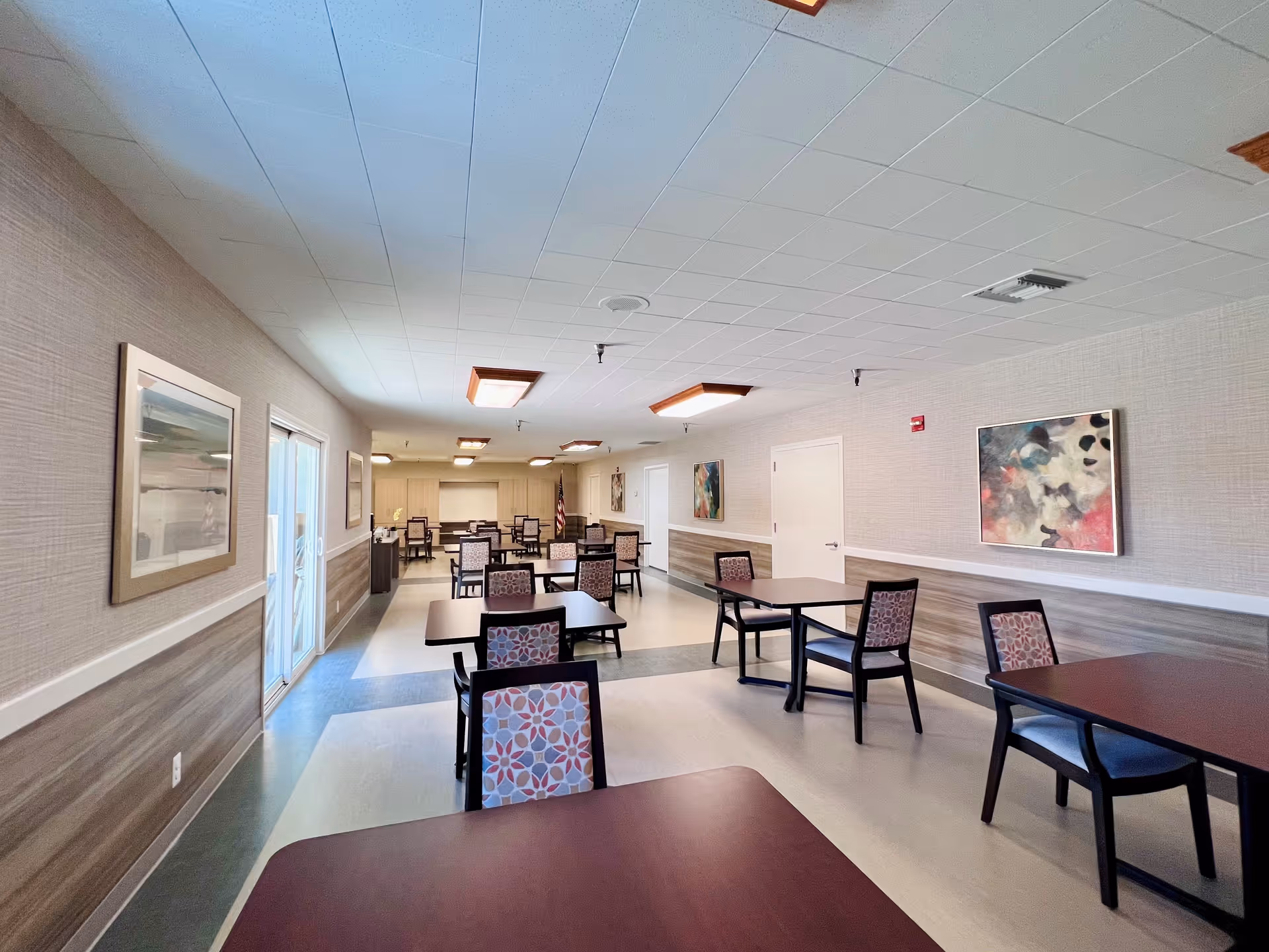 A spacious dining room in a senior living facility with multiple dark wood tables and chairs featuring patterned upholstery. The room has light-colored walls with framed artwork, a tiled ceiling with rectangular light fixtures, and a large window on the left side allowing natural light to enter.