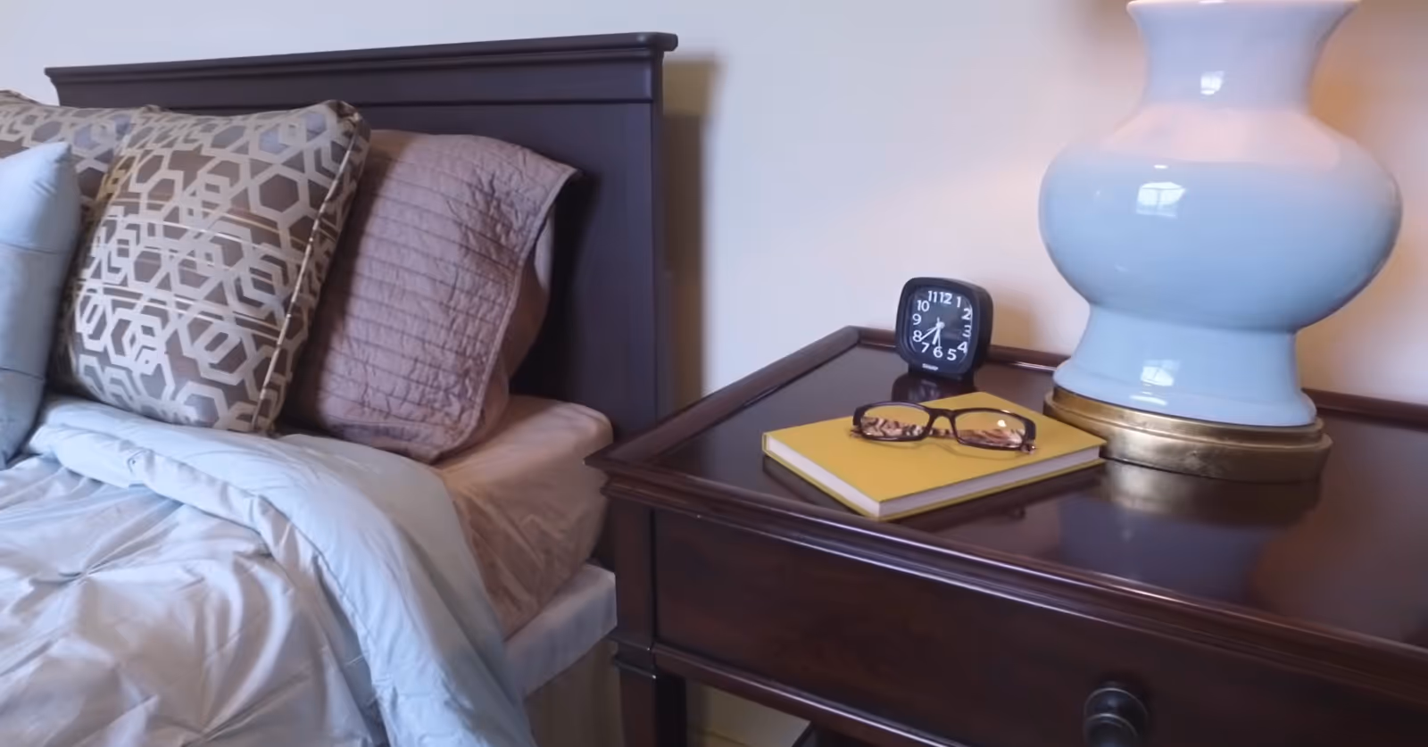 A bedside table holding a lamp, alarm clock, and glasses on a notebook beside a bed with patterned pillows.