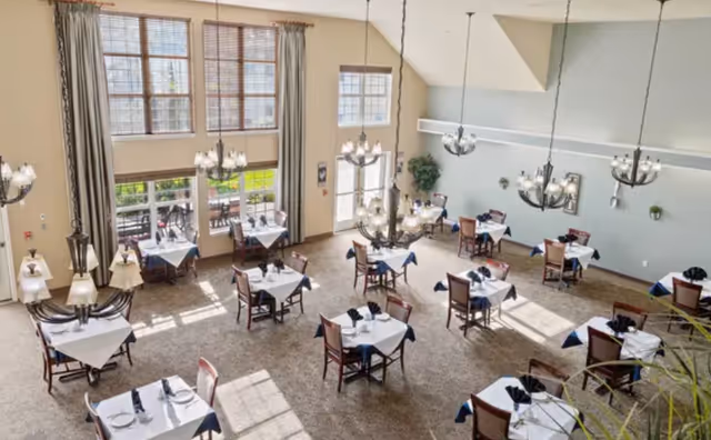 A bright and spacious dining room with multiple tables covered in white tablecloths and blue napkins, surrounded by wooden chairs. Large windows let in natural light, and elegant chandeliers hang from the high ceiling. There is a door leading to an outdoor patio area with more seating.