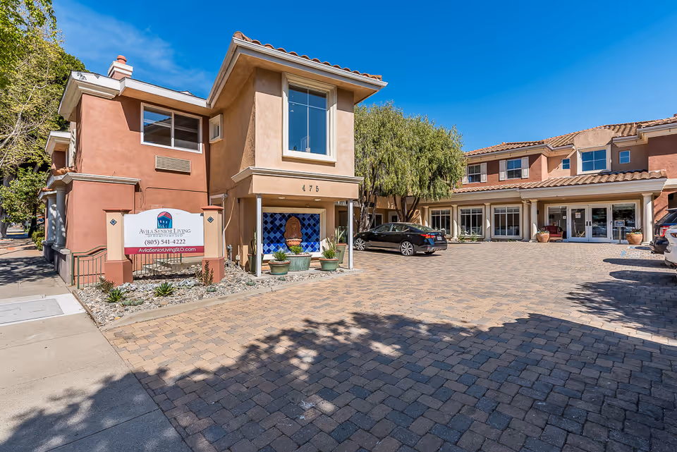 Exterior view of Avila Senior Living At Downtown SLO facility showing a two-story building with a paved driveway and parked cars under a clear blue sky. There is a sign with the facility's name, phone number, and website near the sidewalk.