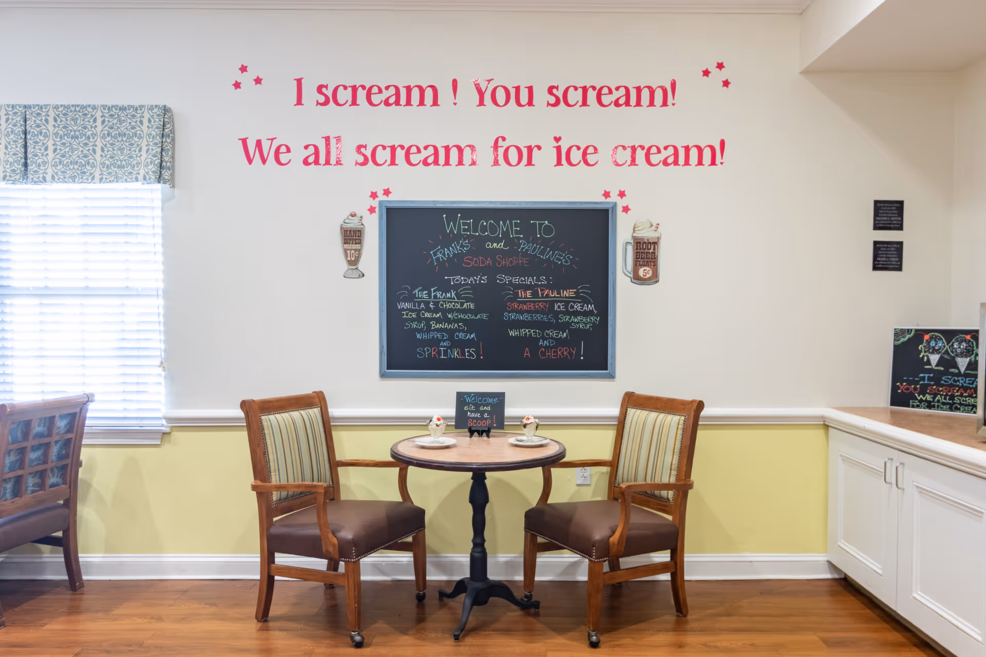 A small dining area with two wooden chairs with striped cushions around a round table set with plates and ice cream desserts. On the wall behind the table is a chalkboard menu with ice cream specials and the text above it reads 'I scream! You scream! We all scream for ice cream!' The room has light yellow and white walls, a window with blinds and a patterned valance, and wooden flooring.