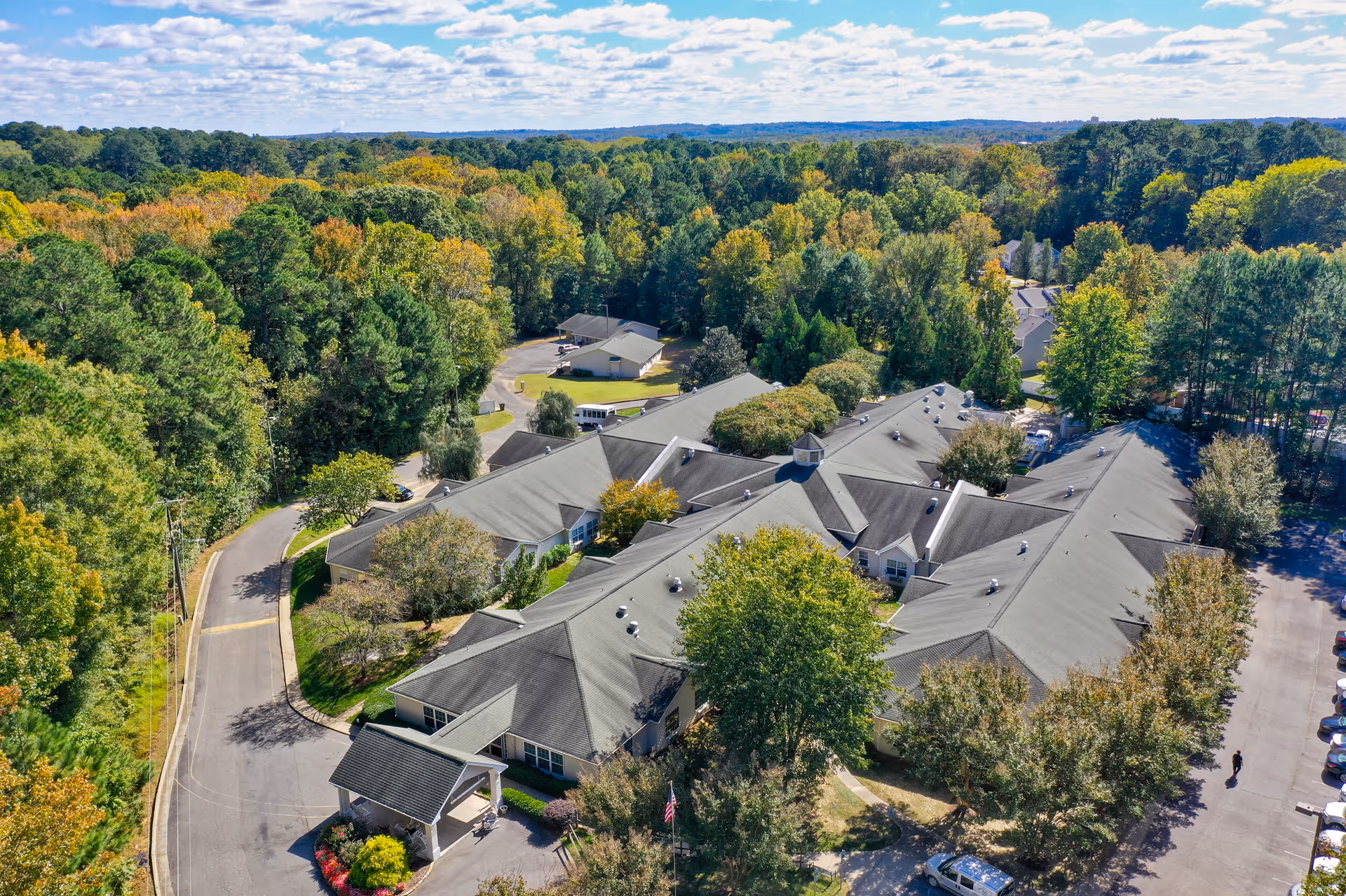 Aerial view of the Cambridge Hills Assisted Living campus surrounded by trees, driveways, and parking areas under a partly cloudy sky.