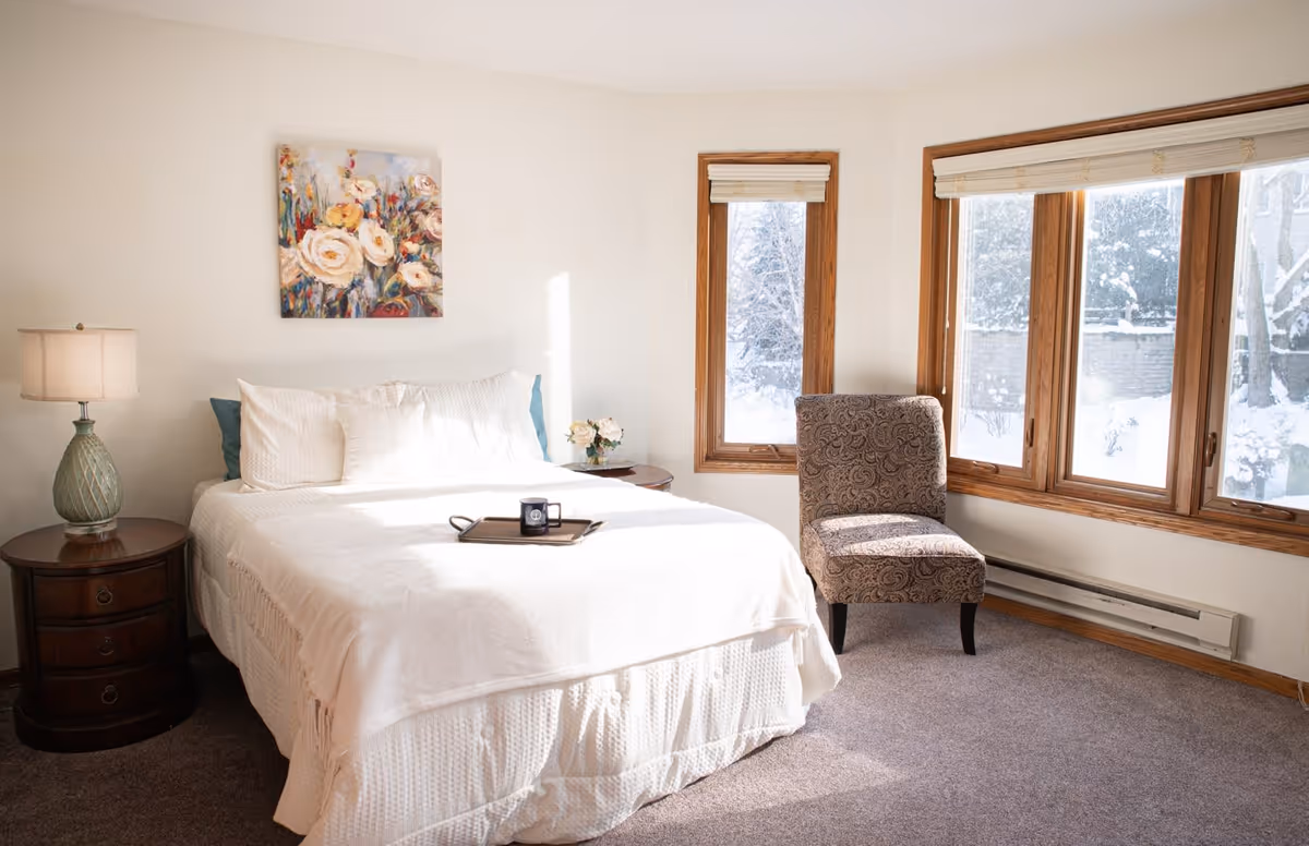 A bright bedroom with a large bed covered in white bedding, a wooden nightstand with a lamp and a small flower arrangement, a patterned upholstered chair, and three large windows showing a snowy outdoor scene.