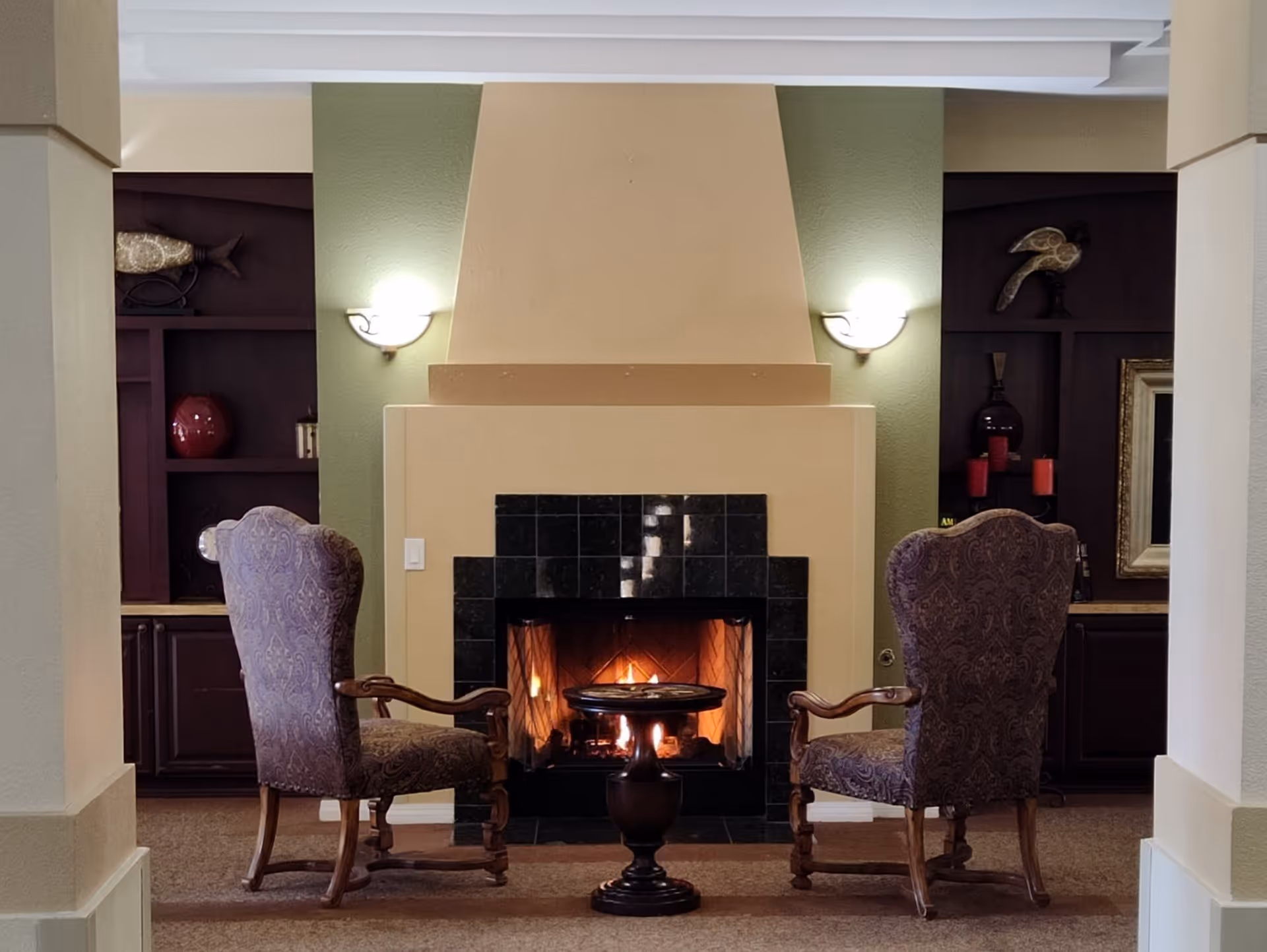 A cozy seating area with two ornate armchairs facing each other across a small round table in front of a lit fireplace with a beige and black tiled surround. The background features built-in dark wood shelves with decorative items and wall sconces providing soft lighting.