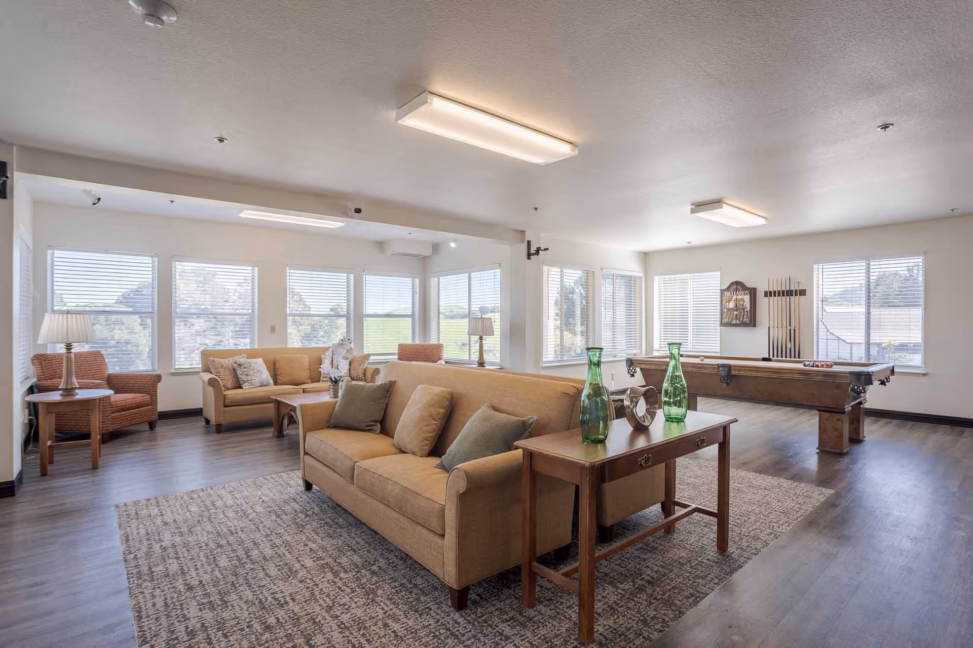 A spacious and bright common living area with large windows letting in natural light. The room features two beige sofas with cushions, a wooden coffee table, a side table with a lamp, and a pool table with cues on the wall. The floor is wooden with a large area rug under the seating area.