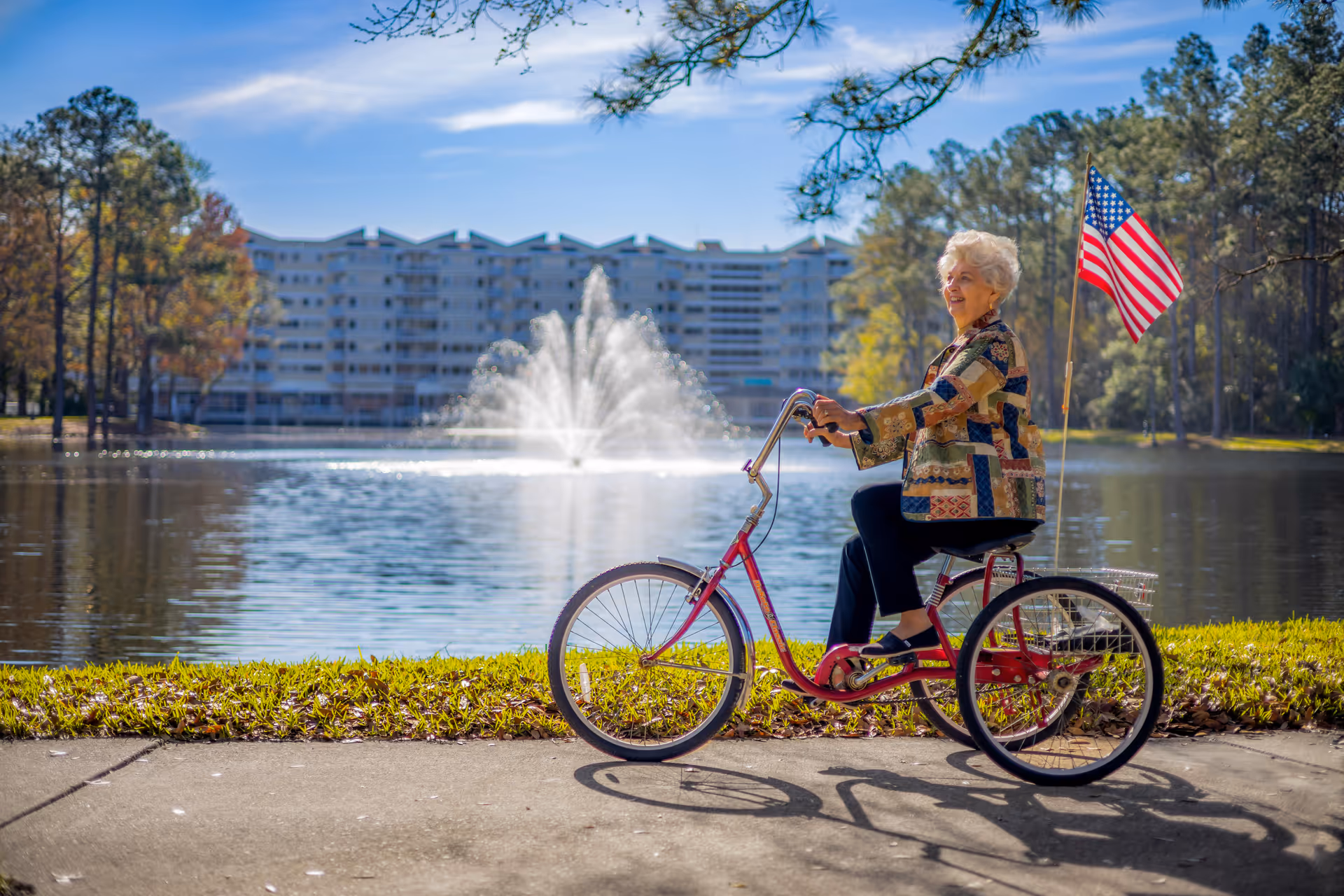 An elderly woman riding a red tricycle with an American flag attached, smiling beside a lake with a large fountain. In the background, there is a multi-story building surrounded by trees under a blue sky.