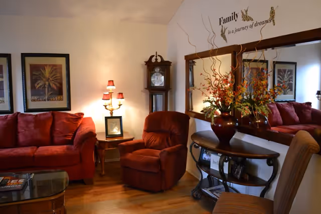 Cozy living room featuring a red sofa and recliner, side table with lamp, grandfather clock, large mirror and console table with a floral arrangement.