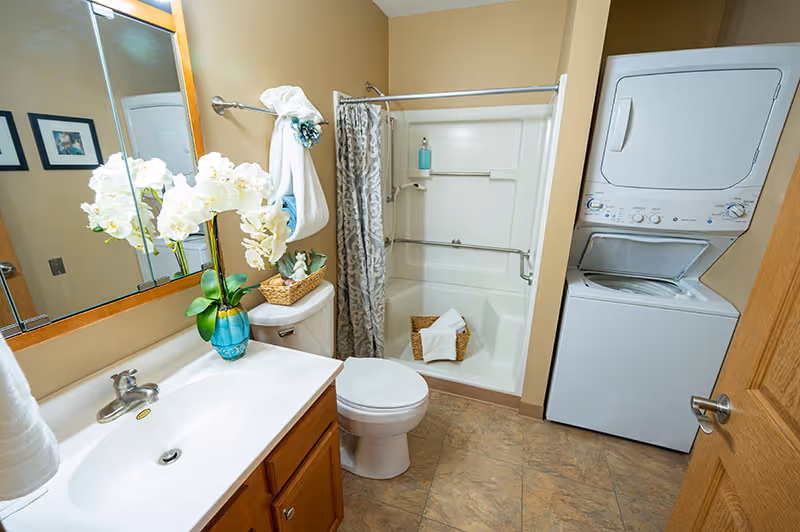 A bathroom with a white sink and wooden cabinet, a toilet, a shower with a patterned curtain, and a stacked washer and dryer unit. There is a vase with white flowers on the sink counter and towels hanging on a rack.