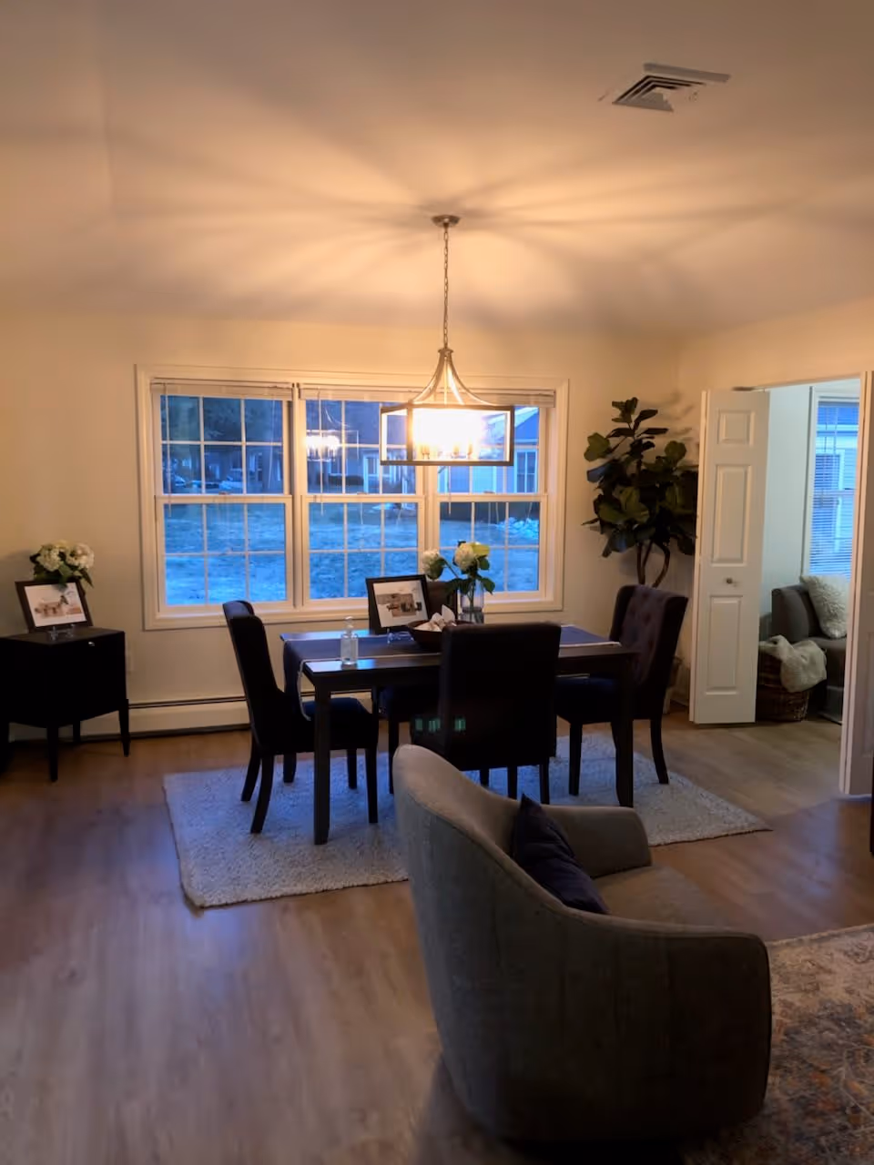 A dining area with a rectangular dark wood table surrounded by four chairs on a light-colored rug. A modern chandelier hangs above the table. There are two framed pictures and flower vases on the table and a small black side table with flowers and a framed picture to the left. A large window shows an outdoor view. To the right, an open doorway leads to a room with a sofa and a plant in the corner.