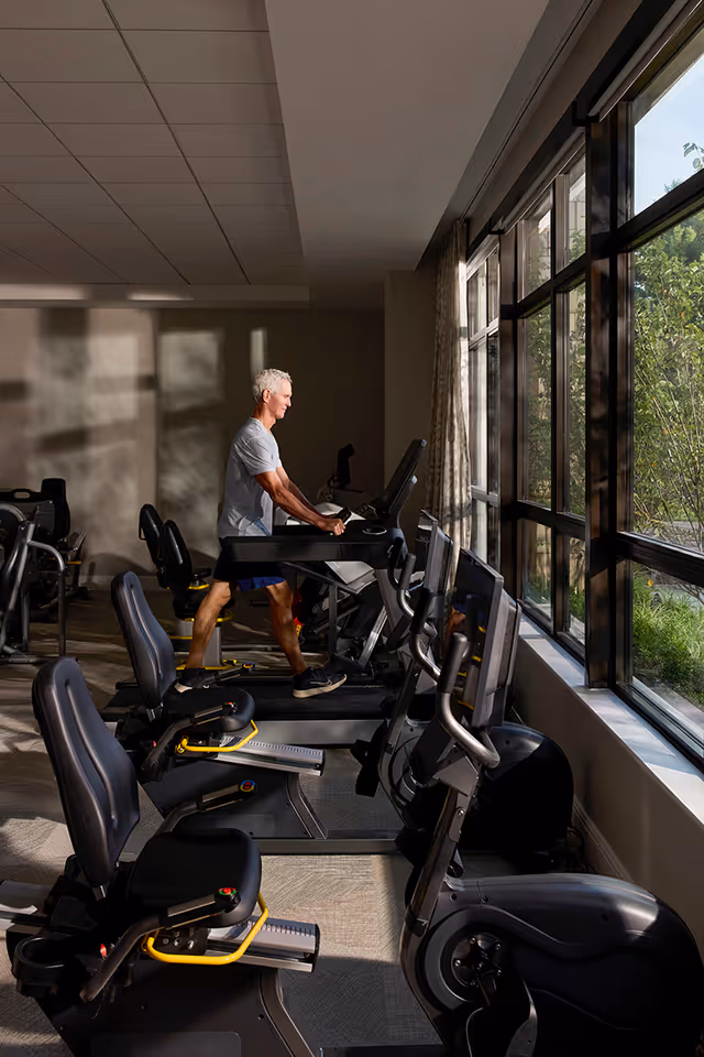 A senior man exercising on a treadmill in a fitness room with large windows letting in natural light and a view of greenery outside. Several other exercise machines are visible in the room.