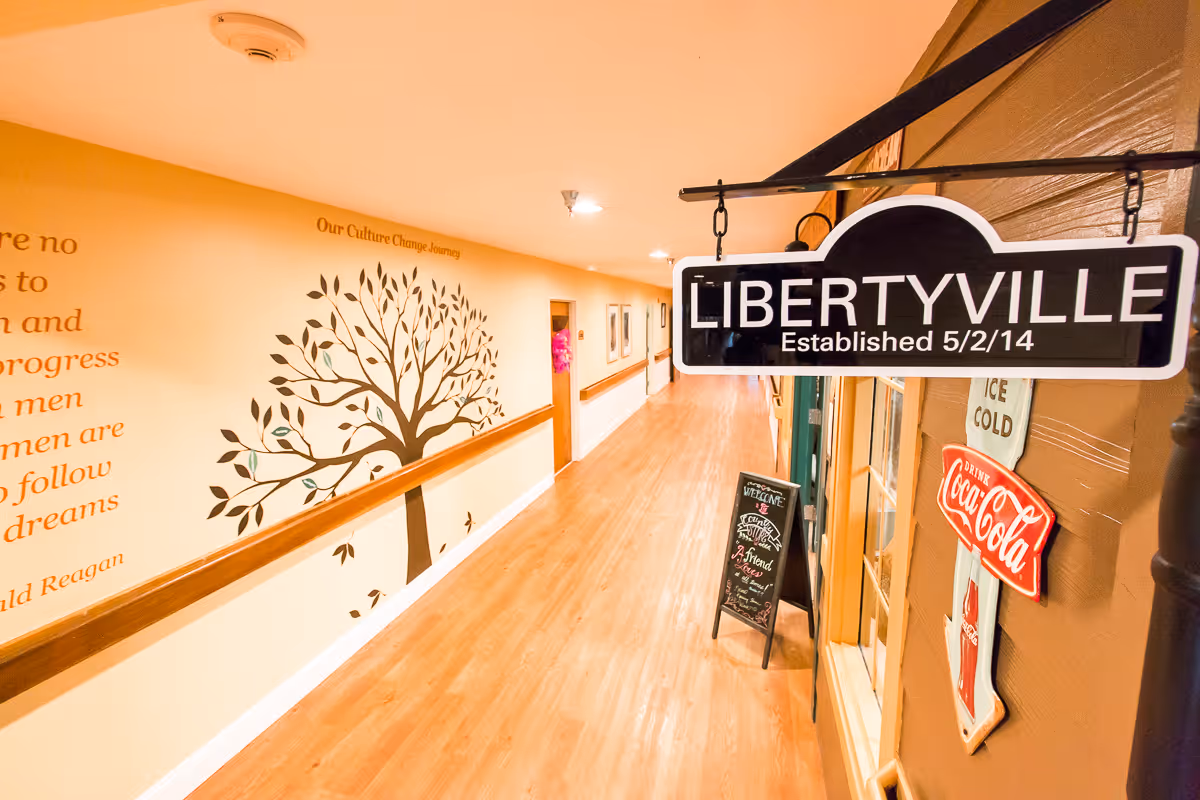 Interior hallway of Liberty Care and Rehabilitation Center featuring a hanging 'LIBERTYVILLE' sign, a tree wall mural, and hardwood flooring.