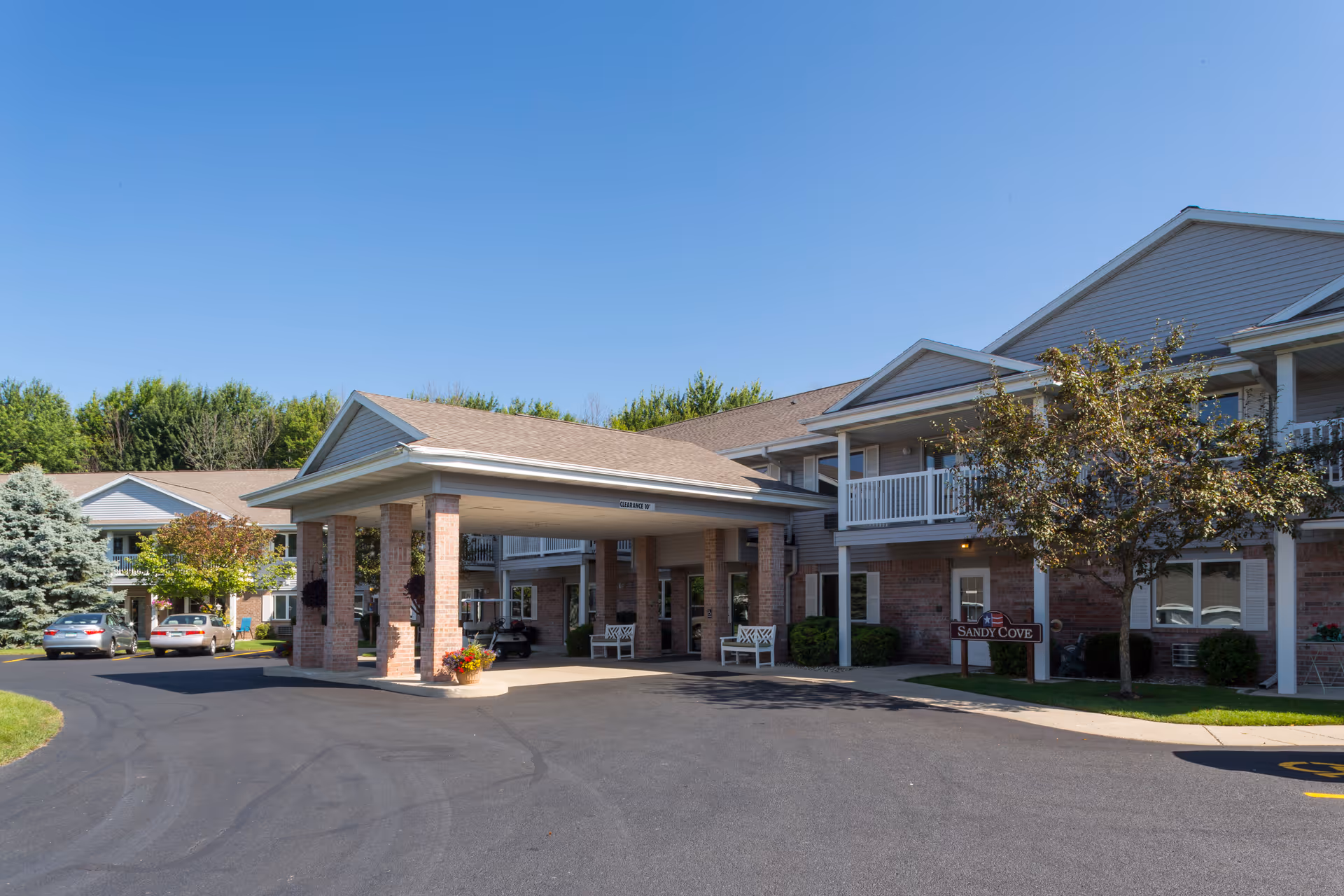 Front exterior of a two-story senior living building with a covered porte-cochere entrance, balconies, landscaping, and parked cars.
