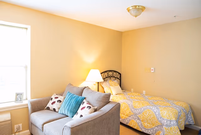 A cozy senior living bedroom with a single bed featuring a yellow and gray patterned comforter, a bedside table with a lamp, and a gray loveseat with decorative pillows near a window.