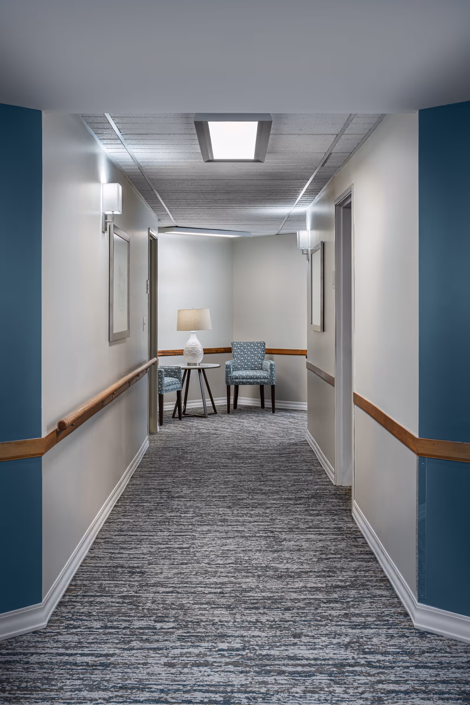 A well-lit hallway in a senior living community with light gray walls, blue accent panels, and a patterned carpet. The hallway features wooden handrails on both sides, two framed pictures on the walls, and two blue patterned armchairs with a small round table and a white lamp at the end.