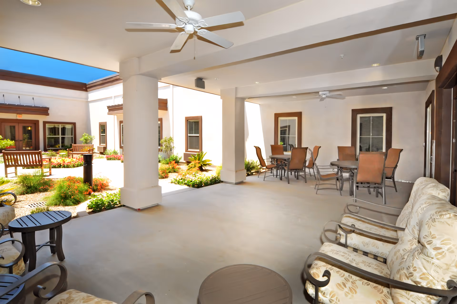 Covered outdoor patio area with ceiling fans, cushioned chairs, round tables with chairs, and a view of a landscaped garden courtyard with benches and plants under a clear blue sky.