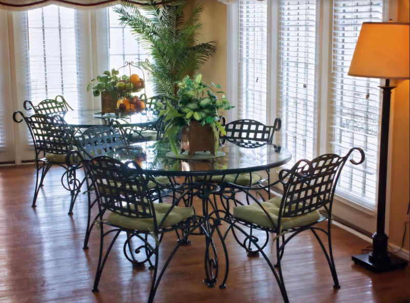 A bright dining area with two round glass tables surrounded by black metal chairs with green cushions. The room has large windows with white blinds letting in natural light. There are green plants on the tables and a tall floor lamp in the corner.