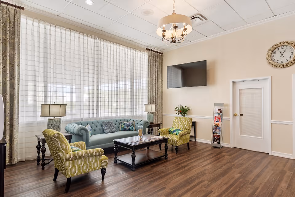 A bright and inviting senior community living room with a large window covered by sheer white curtains and patterned drapes. The room features a light blue tufted sofa with decorative pillows, two green patterned armchairs, a dark wooden coffee table, two side tables with lamps, a wall-mounted flat screen TV, a clock on the wall, and a brochure stand near a white door. The floor is wood with a warm finish, and a chandelier hangs from the ceiling.
